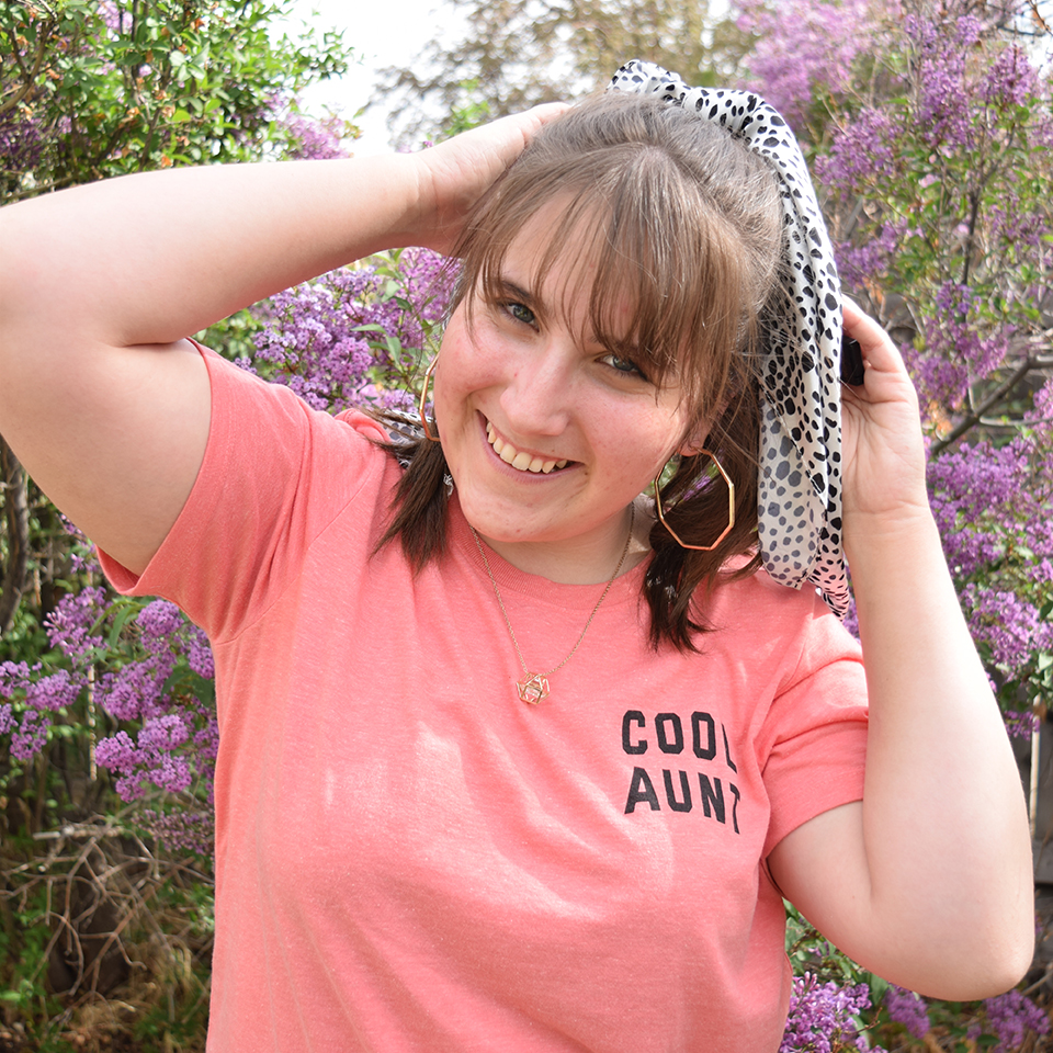 A young woman with brown hair, wearing a pink shirt, standing in front of a purple lilac bush.