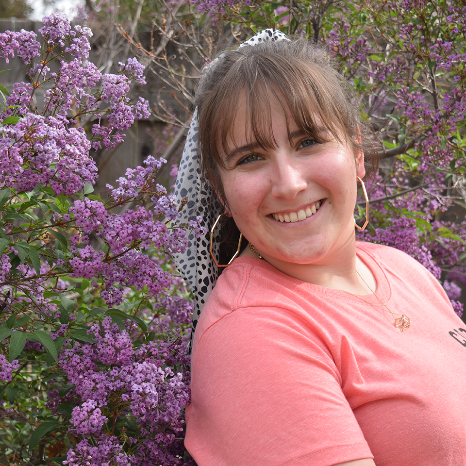 A young woman with brown hair, wearing a pink shirt, standing in front of a purple lilac bush.