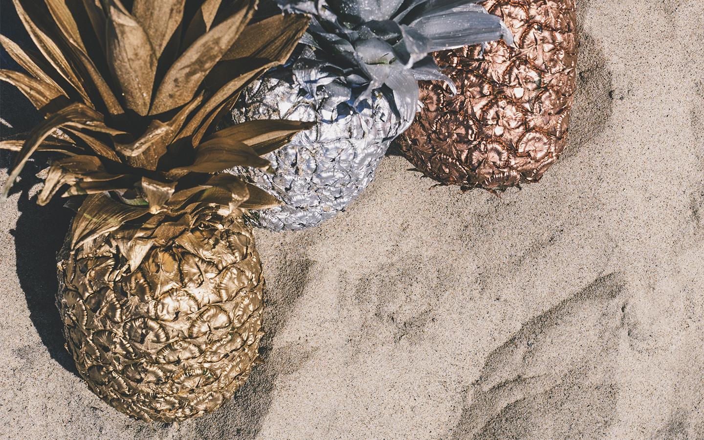 Bronze, copper, and silver painted pineapples sitting on sand.